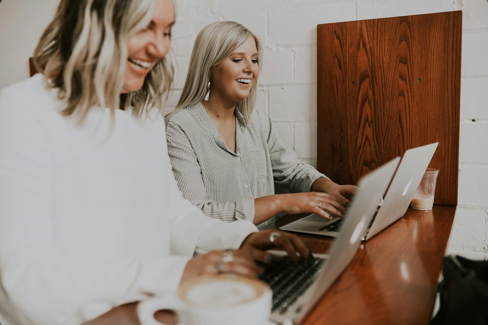 Two women laughing as they work on laptops at a cafe bench