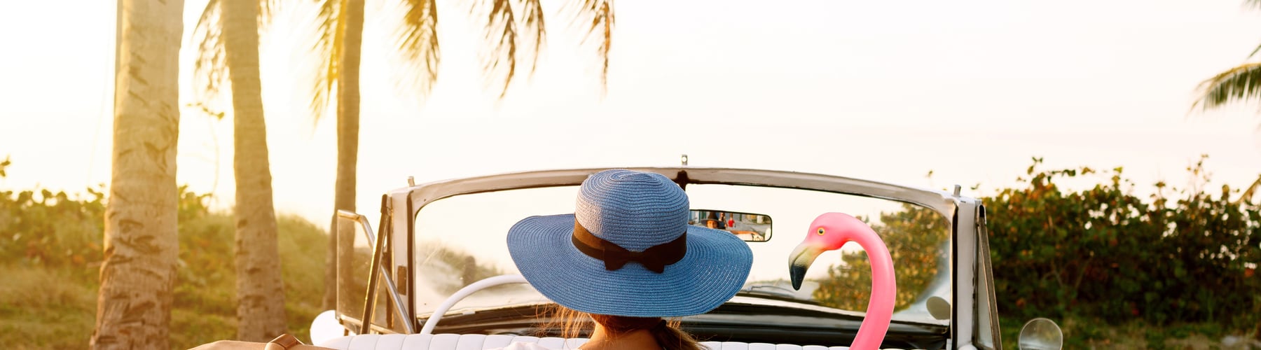 Woman in a sun hat in a white convertible with an inflatable flamingo, under palm trees